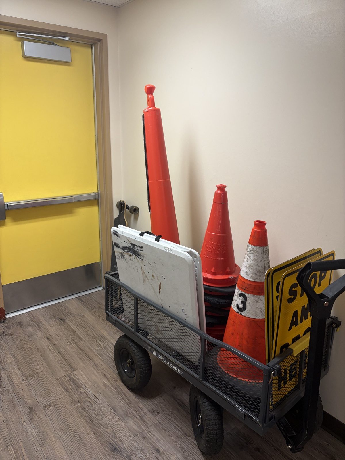 A garden wagon holding traffic cones, folded yellow signs and vests, parked indoors beside a yellow door